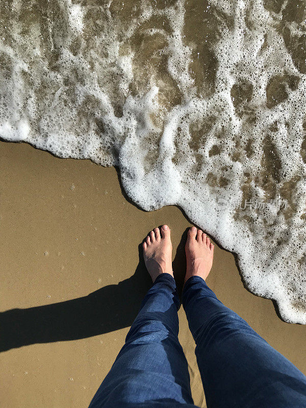 白色的英国男人的脚和紧身的蓝色牛仔牛仔裤在金色的沙滩上被海浪拍打着的海岸线弄湿，用脚趾赤脚划水，夏天的海滨脚印站在海滩上的泡沫/泡沫潮